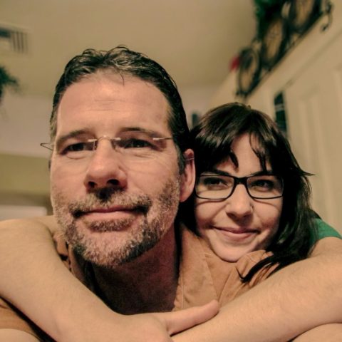 A man and a woman, both in glasses, cheek to cheek; her arms are wrapped around his shoulders.