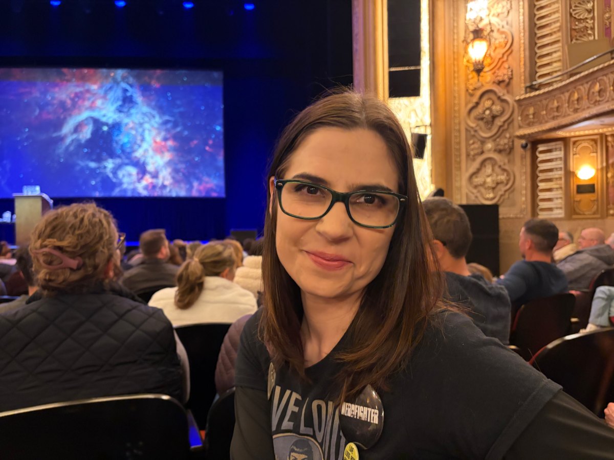 Ash S., smiling and wearing glasses, seated in an audience.